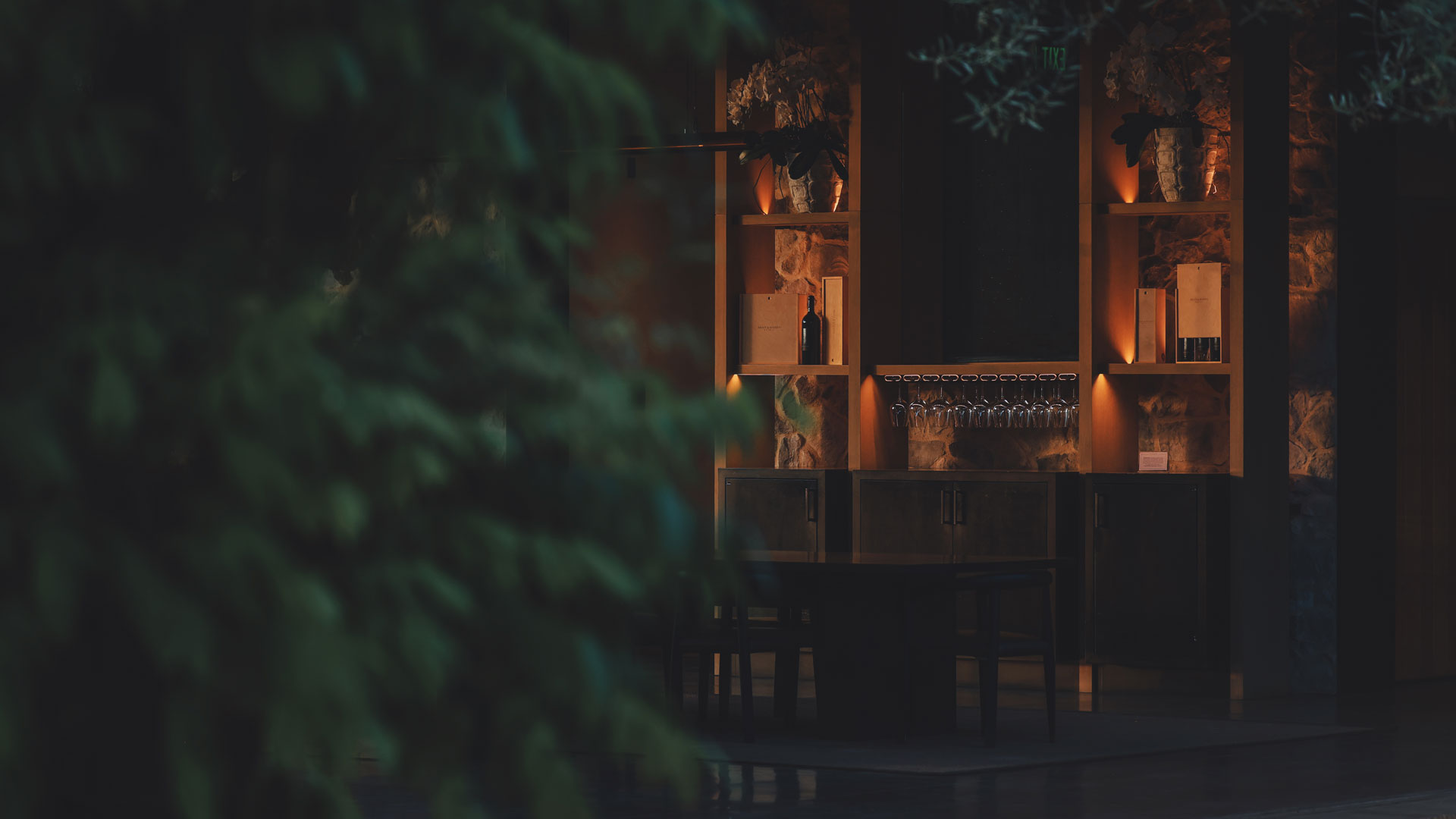 A warm, low-light view of the tasting room at dusk, showcasing a lit-up back bar with wine bottles and glassware.