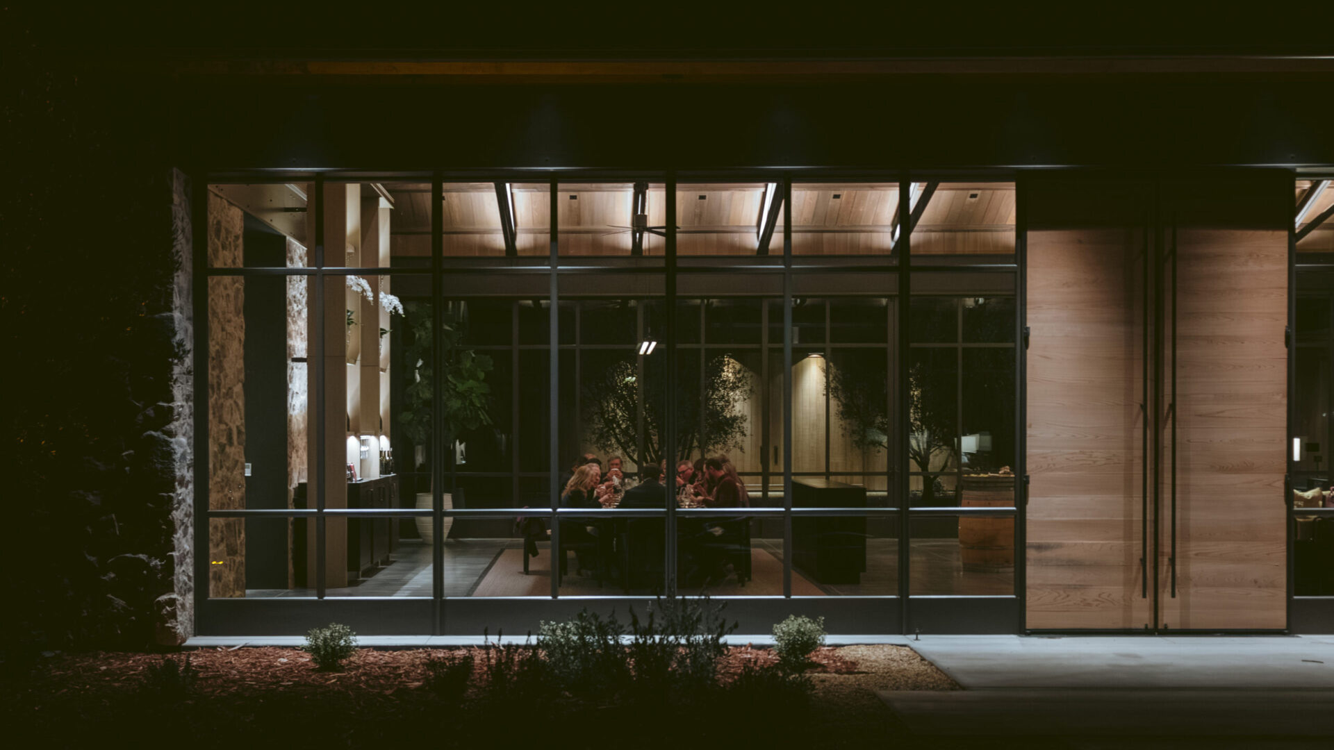 A group of people enjoying dinner at night seen through the large glass doors of the Brave & Maiden tasting room.