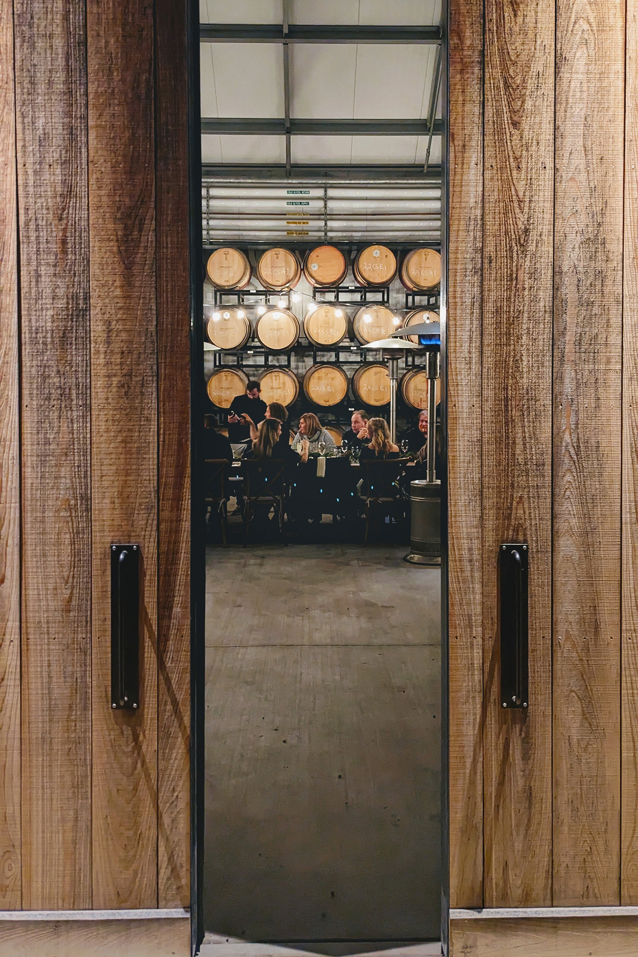 A view through large sliding barn doors into a barrel room where a dinner event is taking place.